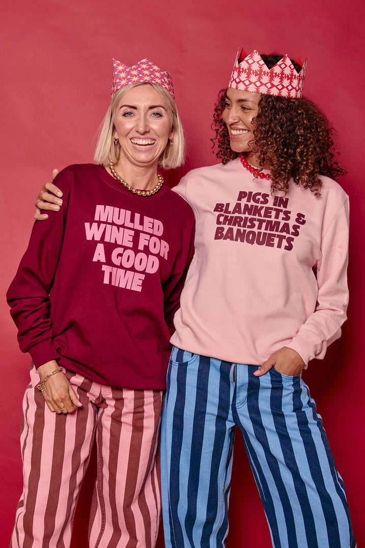 Two women posing together in matching Christmas-themed sweatshirts and striped pants against a red studio backdrop. One sweatshirt is burgundy with white text reading 'MULLED WINE FOR A GOOD TIME' and the other is light pink with dark text reading 'PIGS IN BLANKETS & CHRISTMAS BANQUETS'.