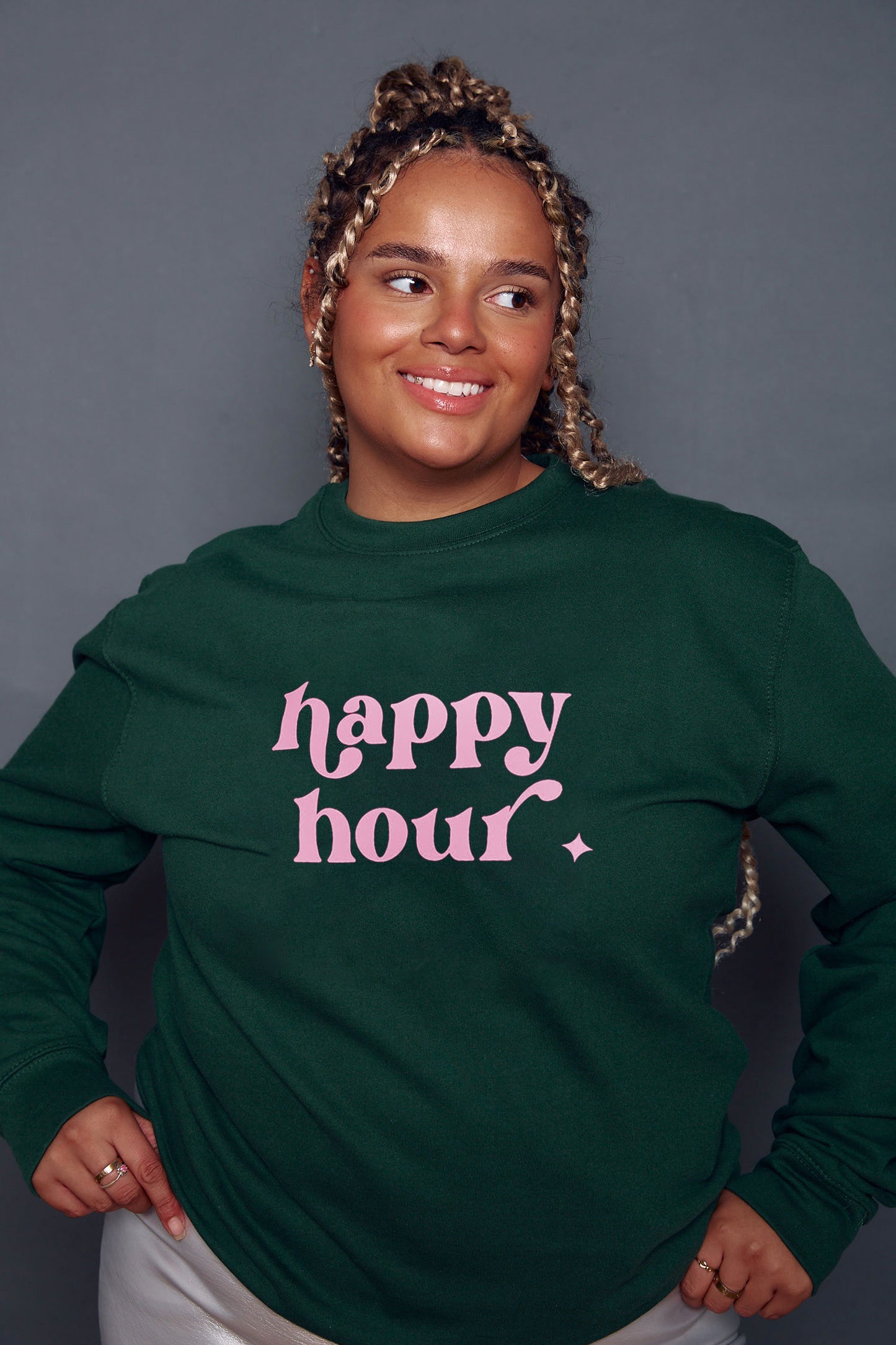 A person wearing a bottle green sweatshirt with the text 'happy hour' in baby pink retro style text.
