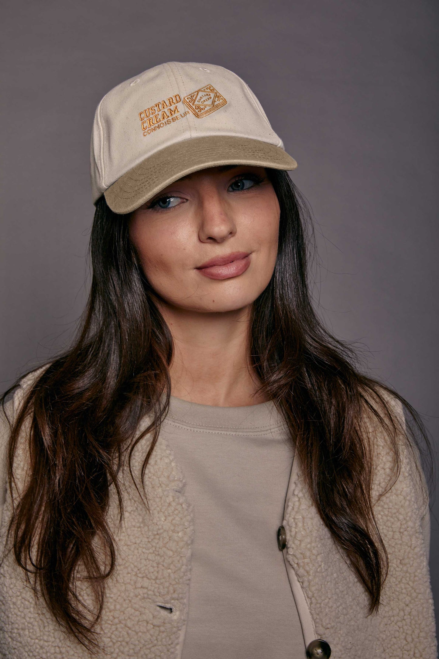 Woman wearing a beige cap with a custard cream design in front of a grey background
