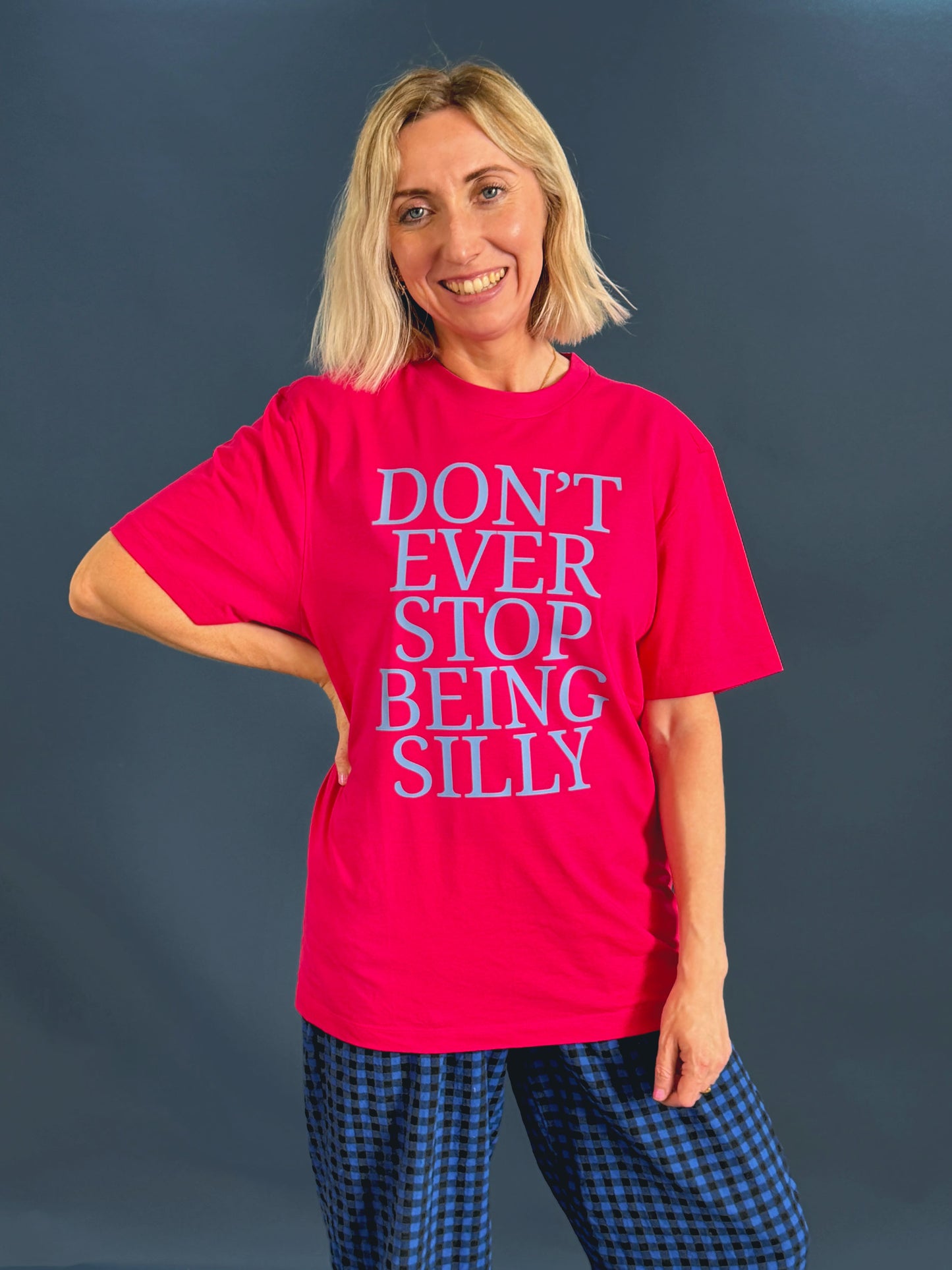 A smiling blonde woman wearing a bright pink graphic t-shirt with the text 'DON'T EVER STOP BEING SILLY' in light blue and dark pink from Rock On Ruby, paired with blue and black checkered trousers, against a dark gray studio background.
