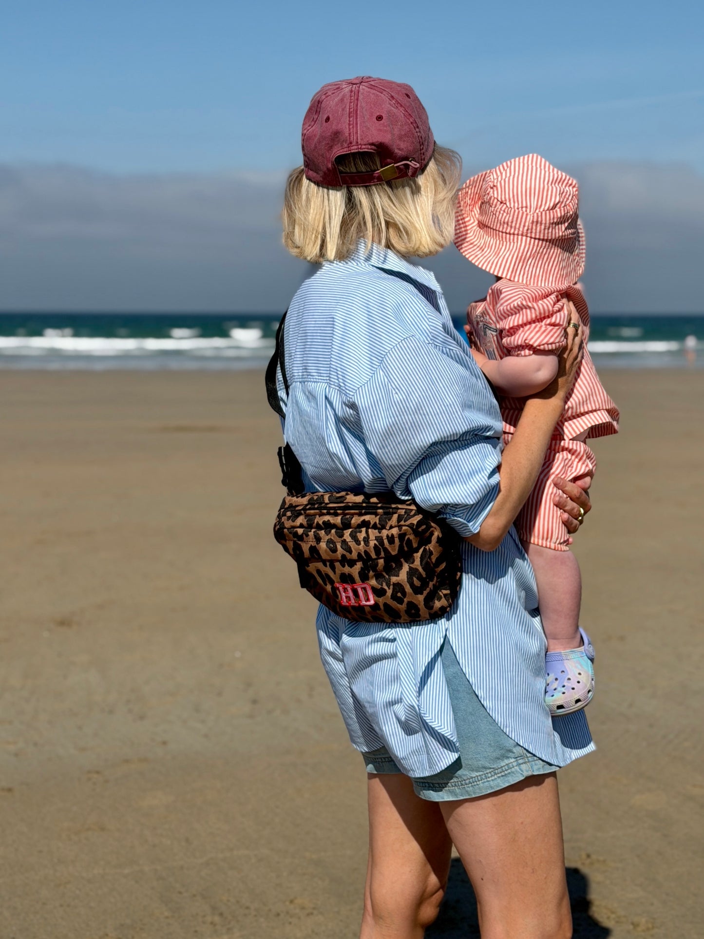 A mum and child standing on a sandy beach, viewed from behind. The woman wears a light blue striped shirt, denim shorts, a dusty rose cap, and carries a leopard-print crossbody bag with pink 'HD' lettering.