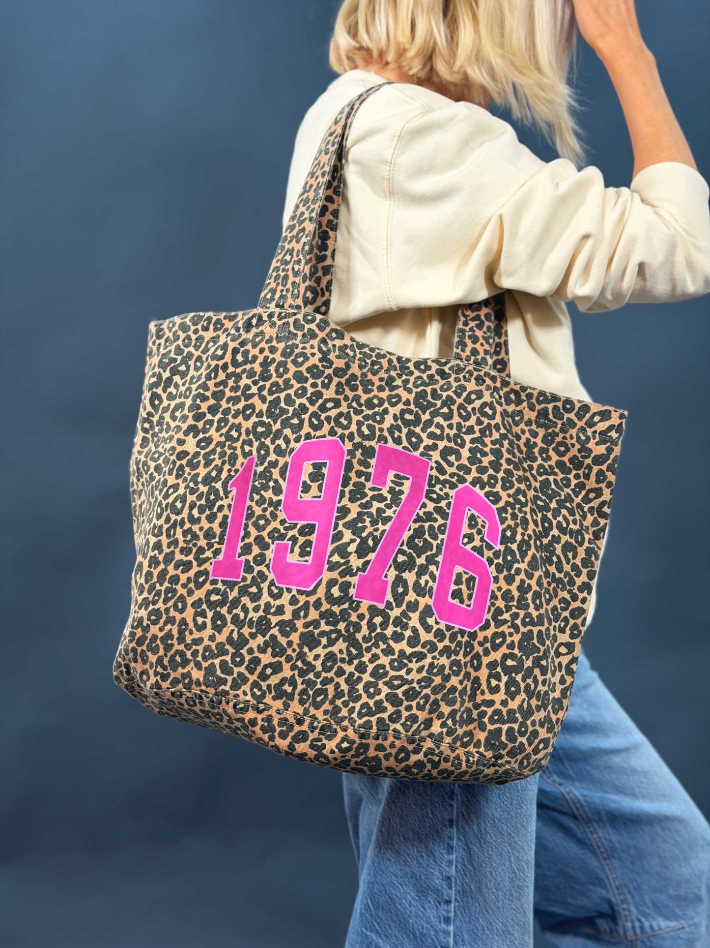 Leopard print tote bag with '1976' in pink being carried by a person against a blue background