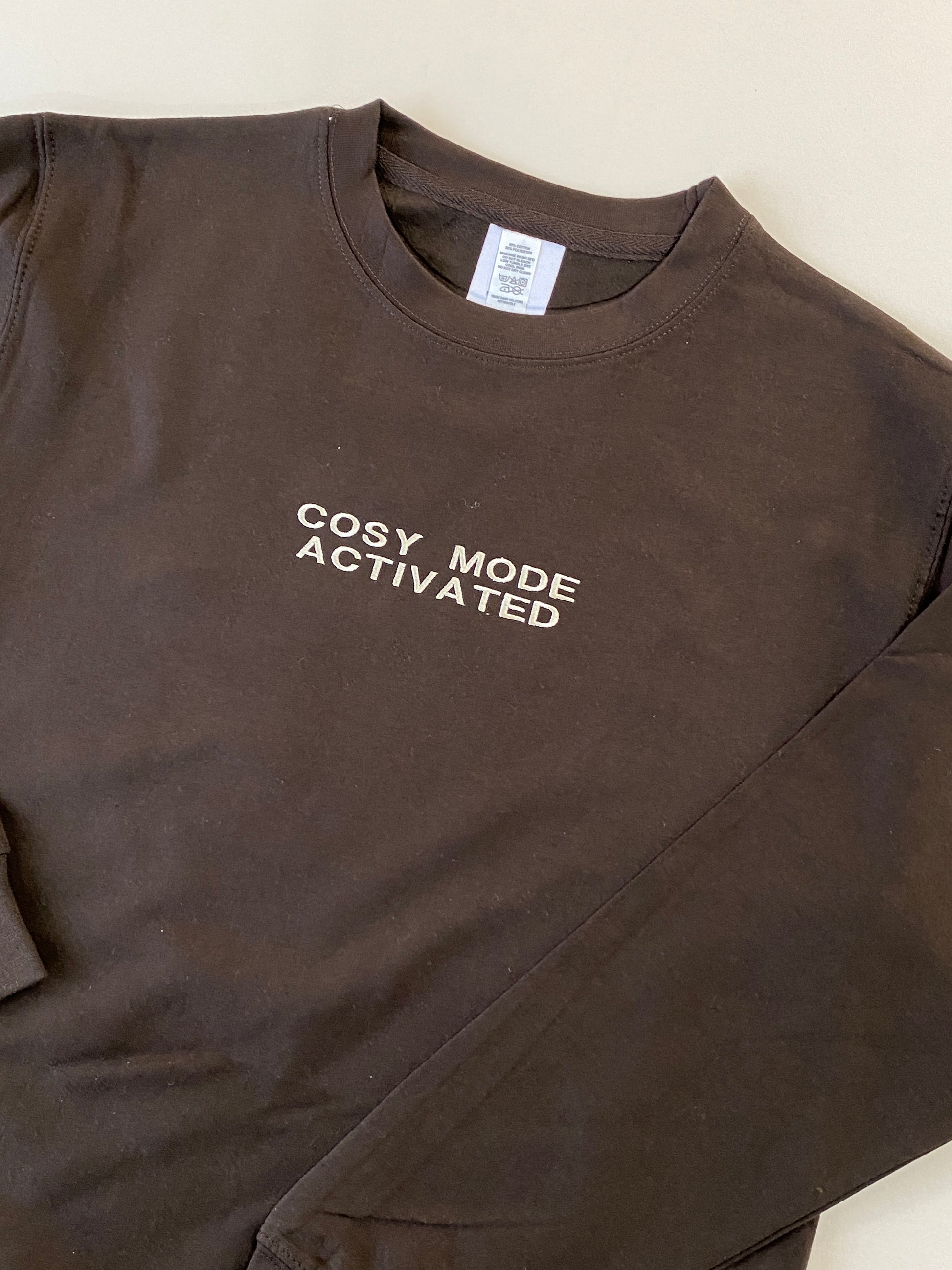 Brown sweatshirt with 'Cosy Mode Activated' embroidered text on a beige background