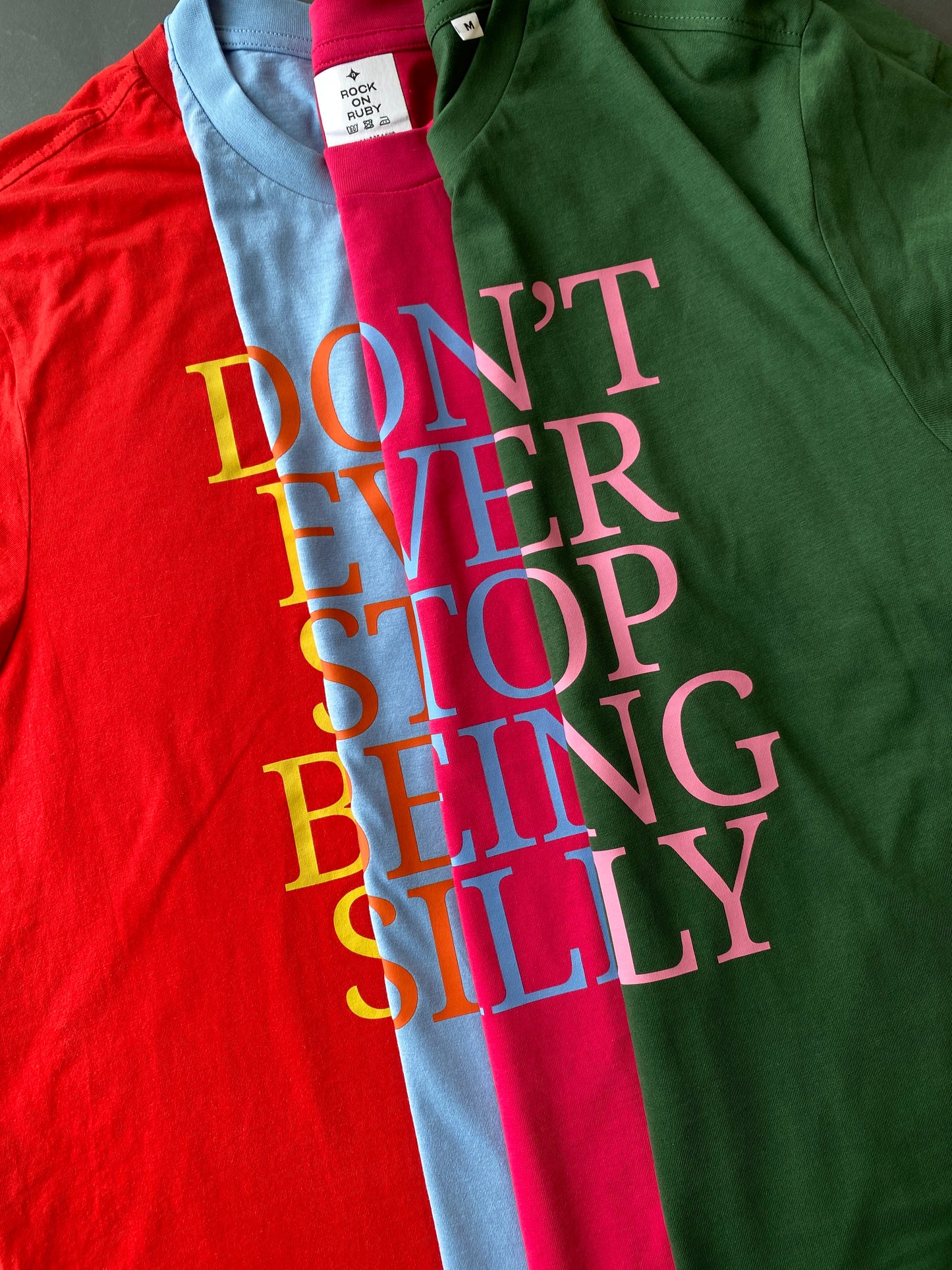A close-up of graphic t-shirts folded together featuring the text 'DON'T EVER STOP BEING SILLY' in large, letters. The shirts are shown going from red on the left, to light blue, hot pink and dark green on the right.