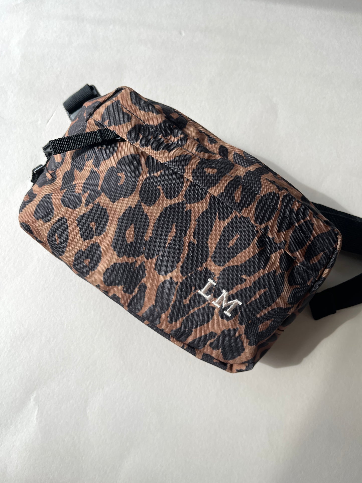 A small leopard-print crossbody bag with a brown and black pattern, featuring a cream 'LM' monogram on the front and black straps, laid flat on a white surface.