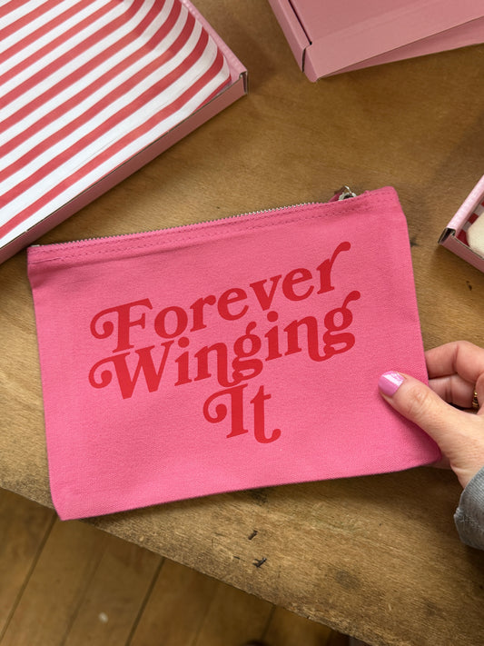 Pink pouch makeup bag with 'Forever Winging It' text held by a hand on a wooden surface