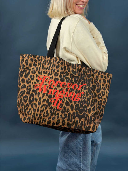 Person holding a leopard print tote bag with red text slogan 'forever winging it' against a dark background