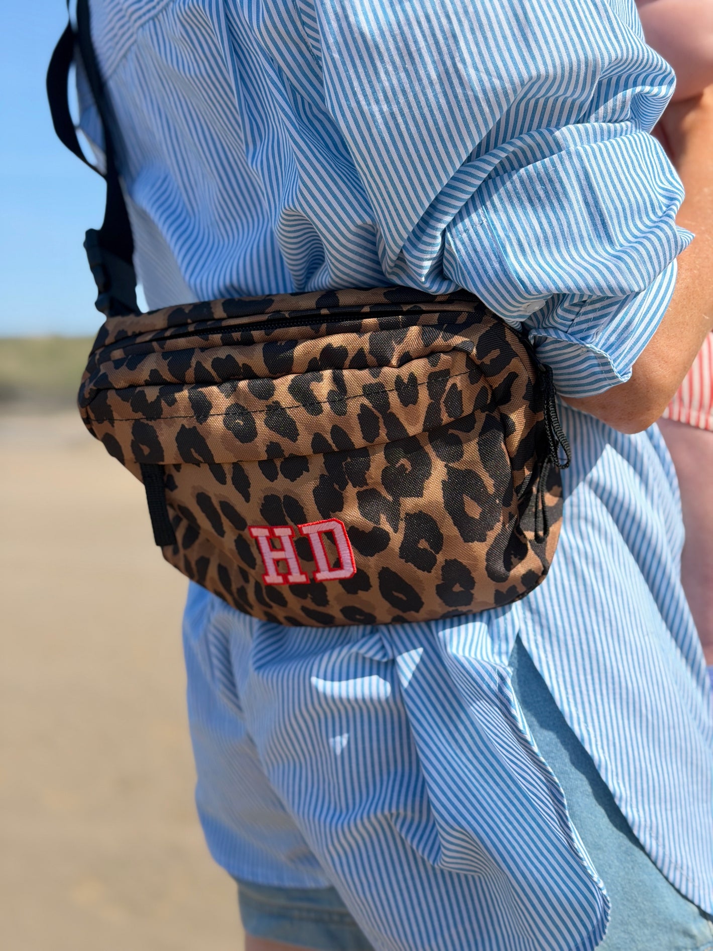 A leopard print crossbody pack with a pink embroidered 'HD' monogram, worn by a person in a blue and white striped shirt.