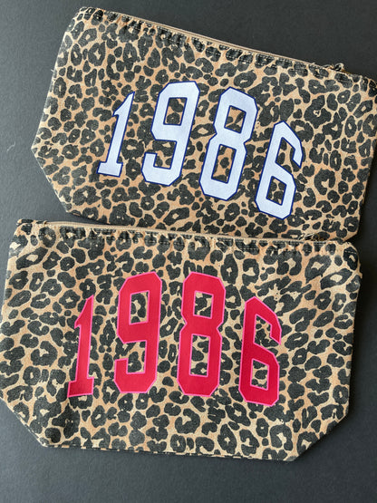Two leopard print makeup bags with '1986' printed on them, one in off white and one in hot pink, against a dark background.
