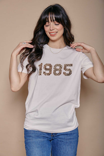 Woman wearing a cream t-shirt with '1985' printed on it in leopard print stood against a beige background