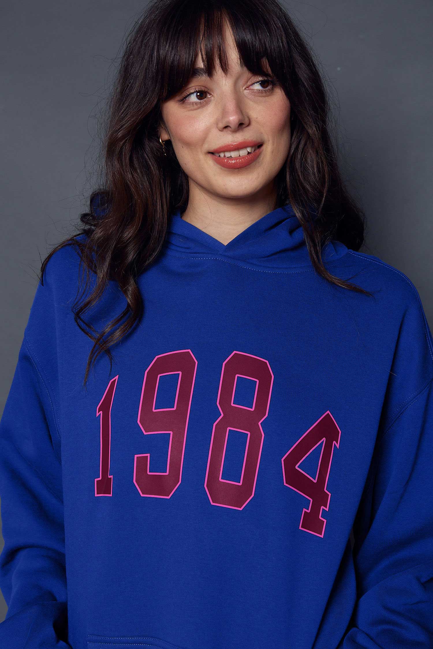Person wearing a blue hoodie with '1984' printed on it in burgundy and pink against a grey background