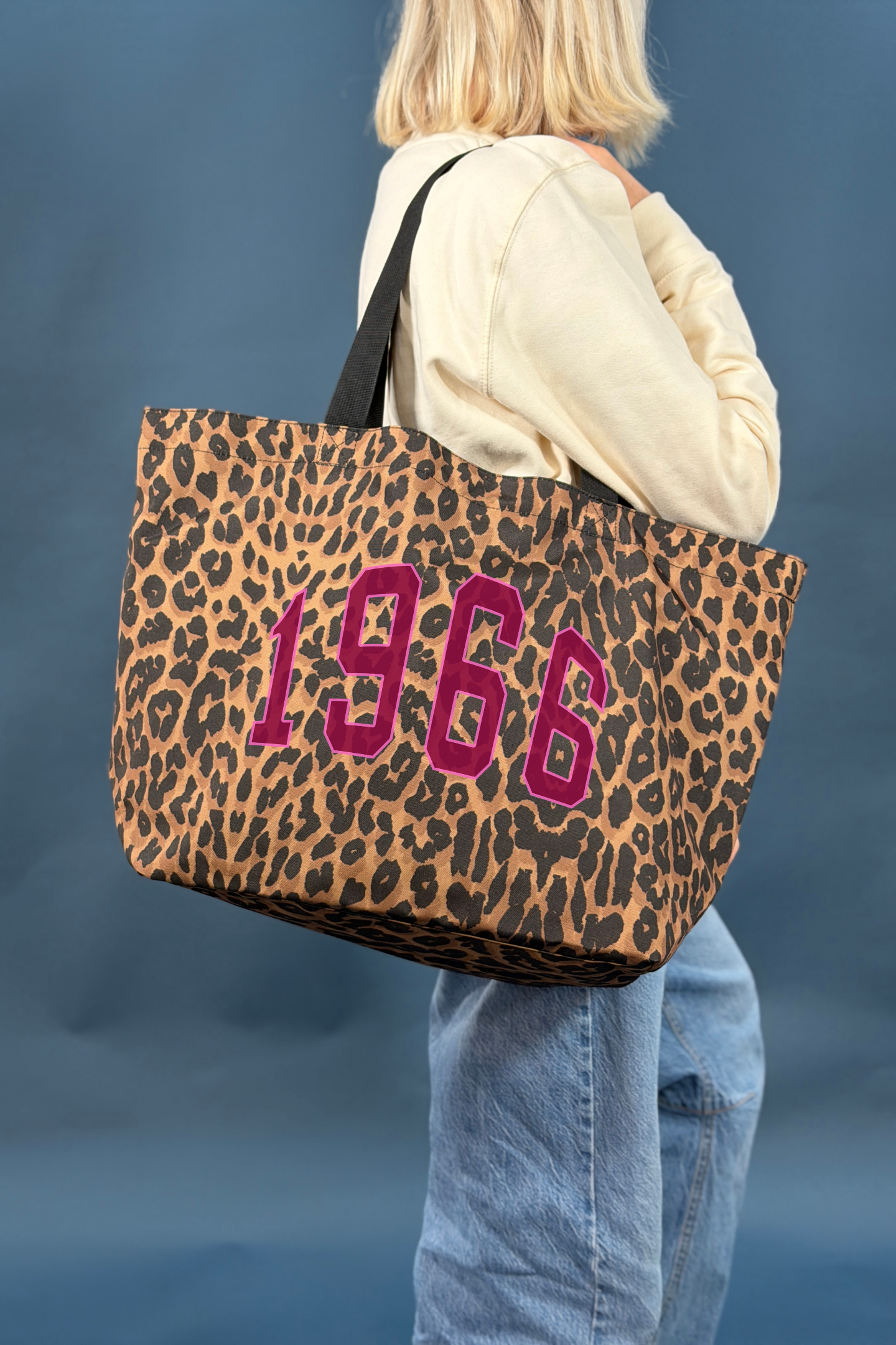Person holding a leopard print tote bag with '1966' in burgundy text against a blue background