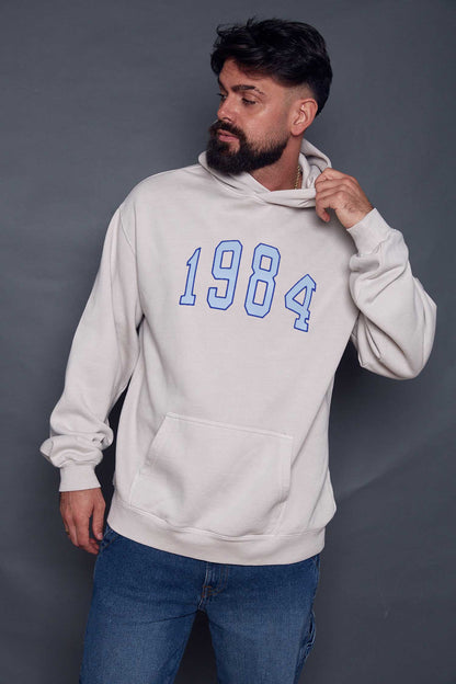 Man wearing a cream hoodie with '1984' printed in blue stood in front of a dark grey background