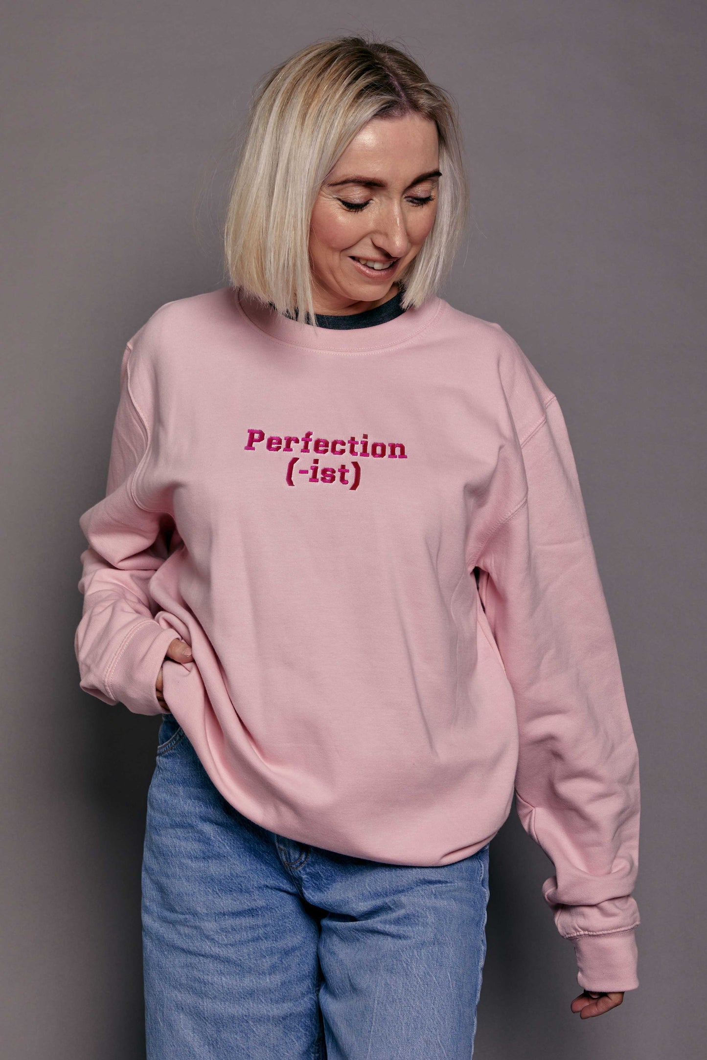 Person wearing a baby pink sweatshirt with hot pink embroidered text, standing against a gray background