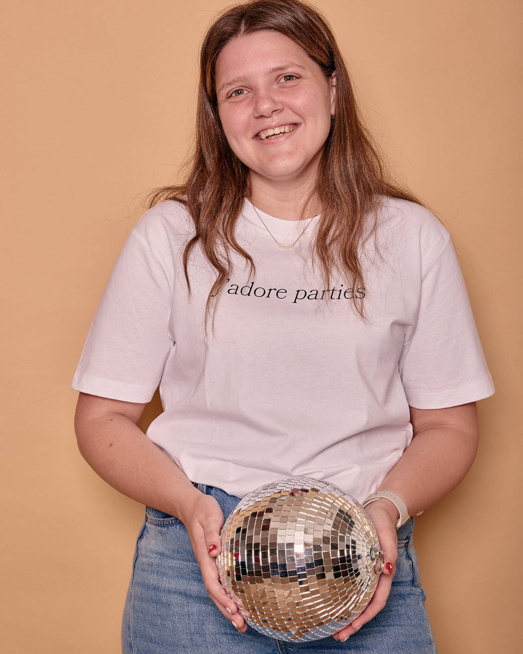 A smiling woman with long light brown hair stands against a warm beige studio background, wearing a white graphic T-shirt with the words 'j'adore parties' and blue jeans. She is holding a mirrored disco ball.