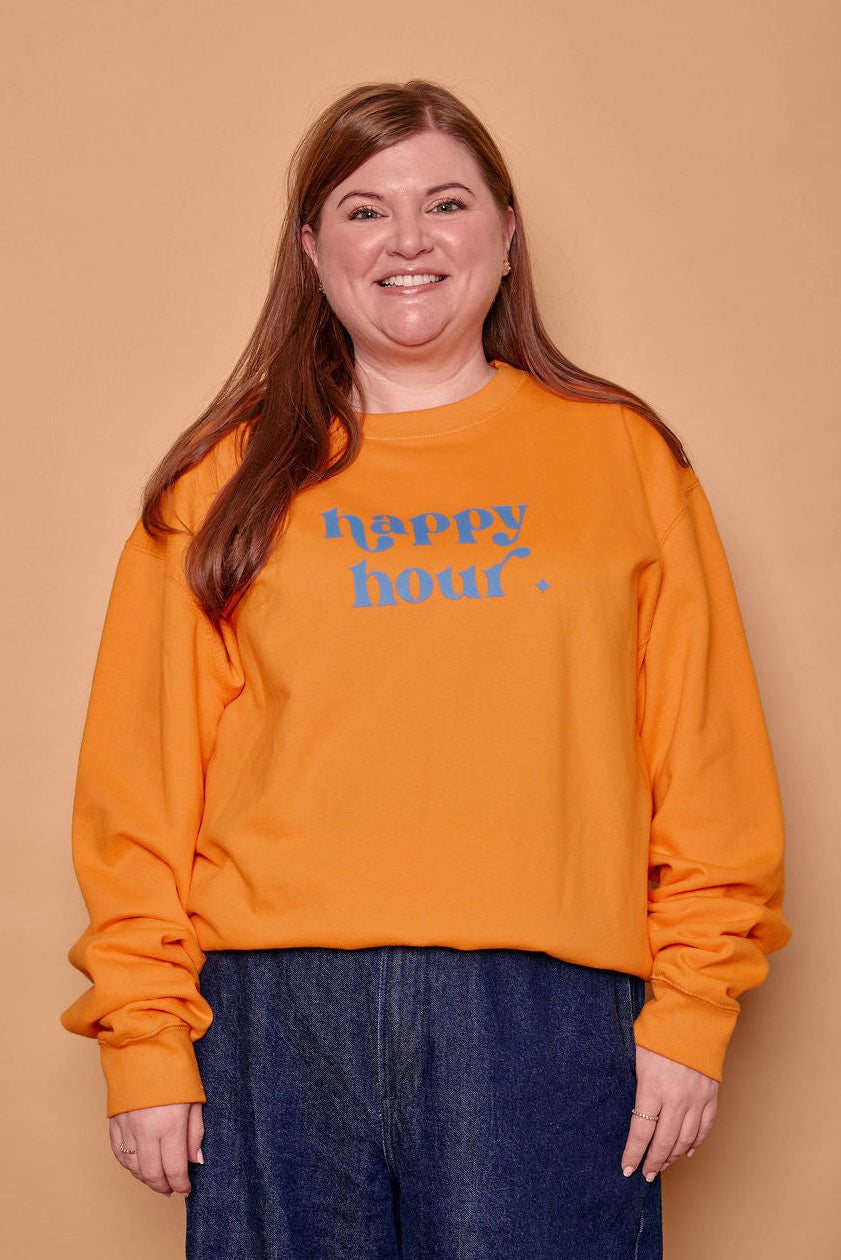 A smiling woman with long light brown hair stands against a solid tan background, wearing a bright orange crewneck sweatshirt with 'happy hour.' printed in blue across the chest, paired with dark blue wide-leg jeans.