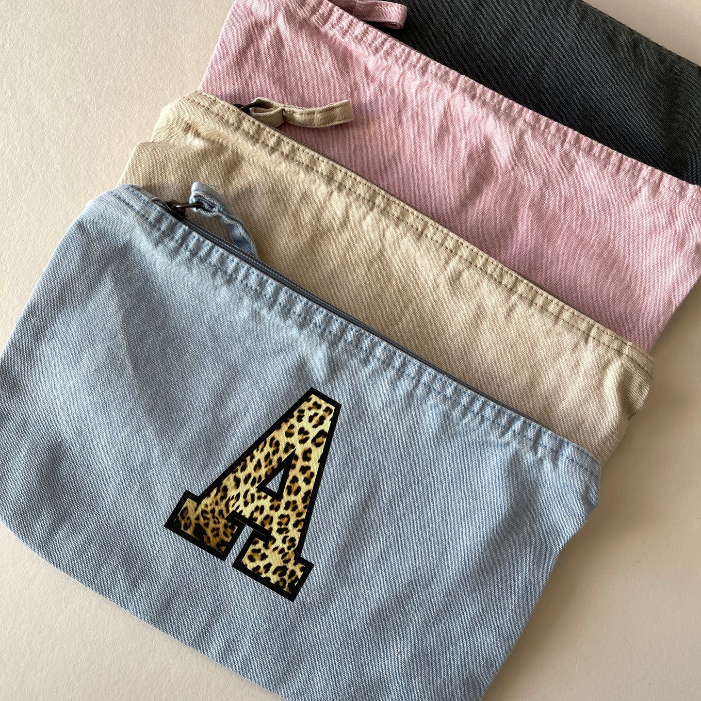 Four fabric pouches in different colors with a leopard print letter 'A' on a beige background.