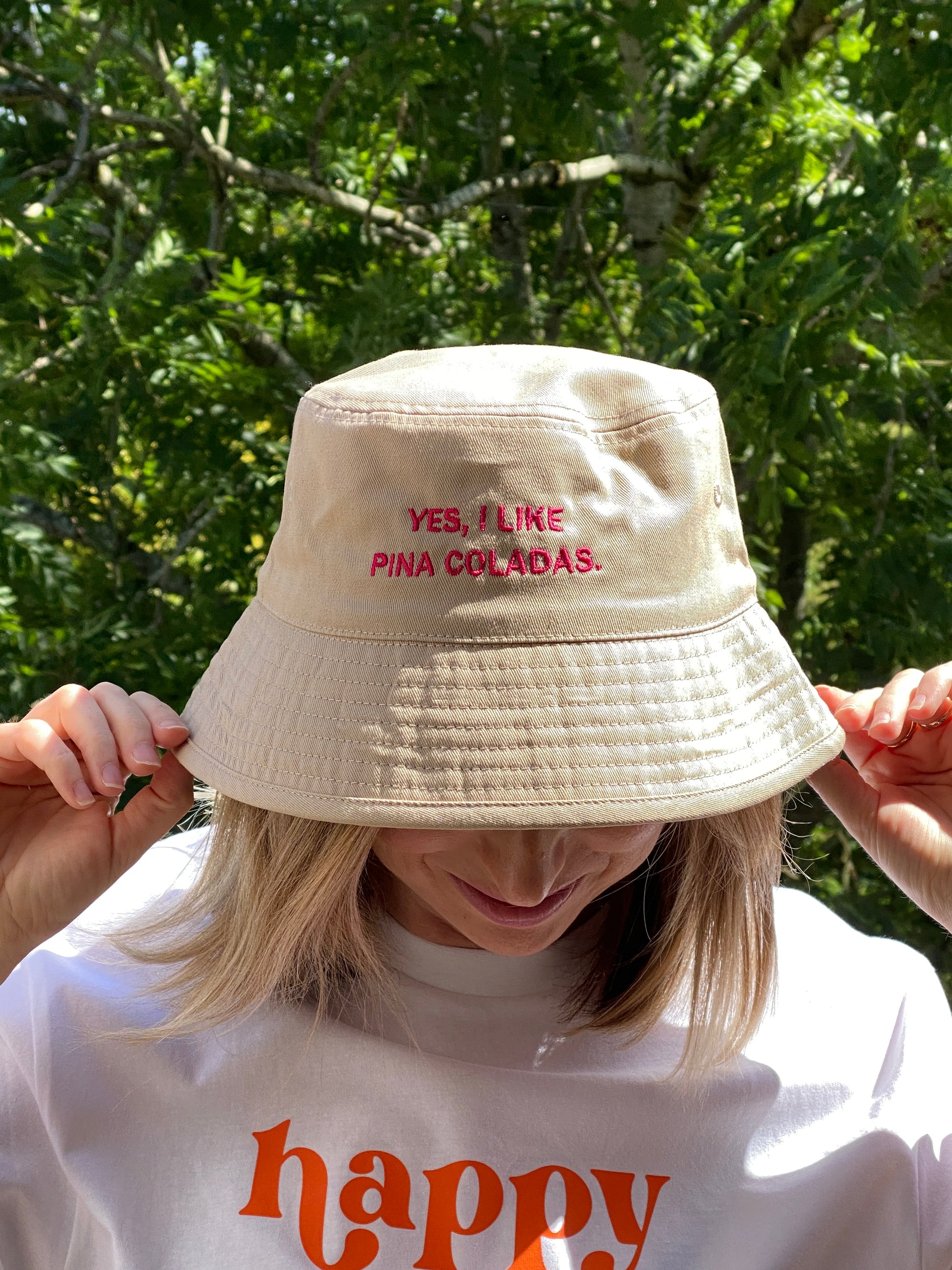 Person wearing a beige bucket hat with hot pink text 'Yes I like pina coladas' and a white shirt with orange text, outdoors.