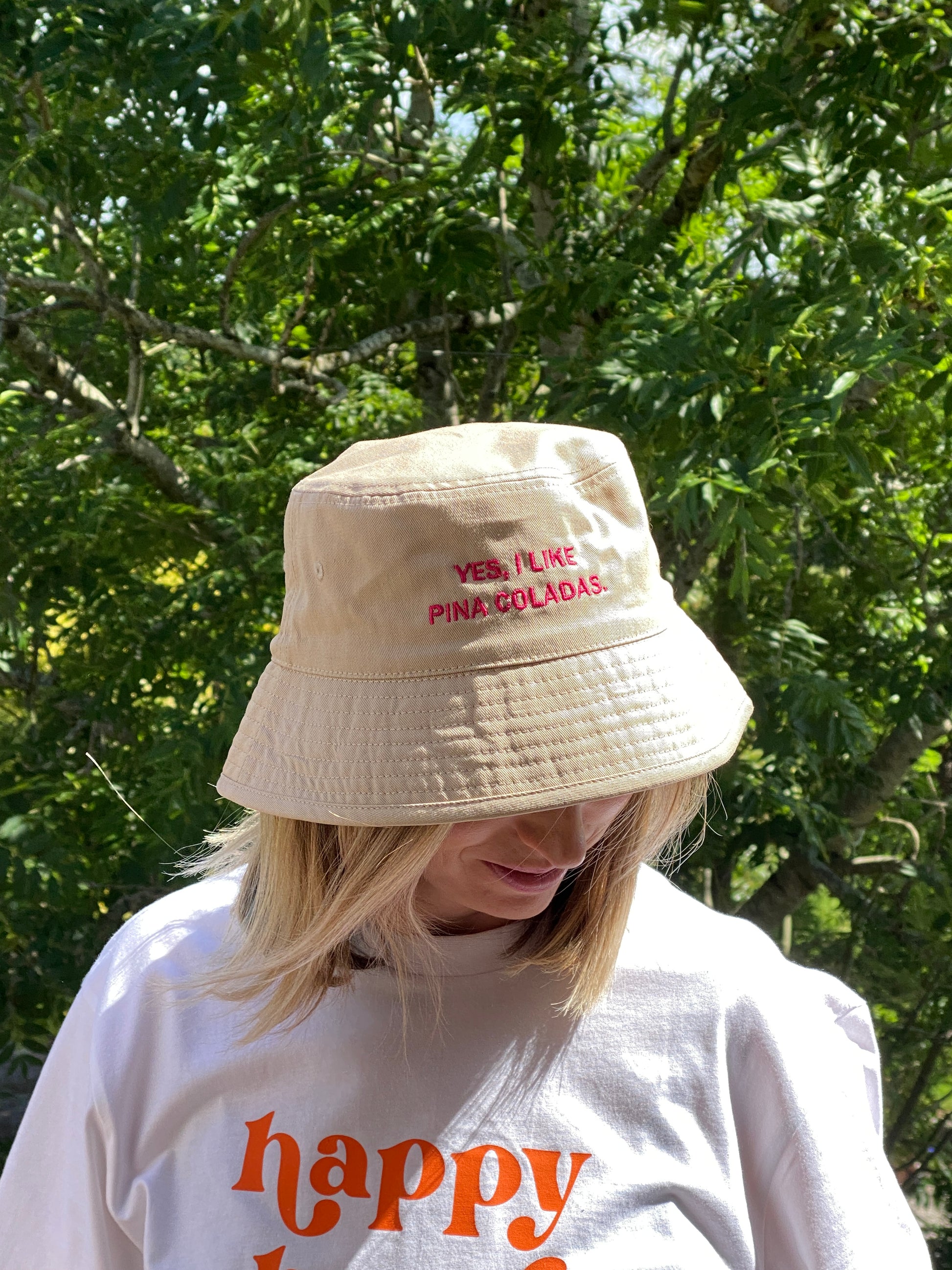 Person wearing a beige bucket hat with text 'yes I like pina coladas' and a white t shirt with 'happy hour' printed on it, standing in front of green foliage.