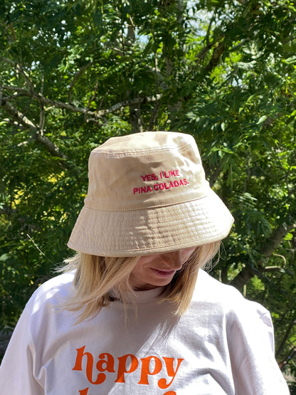 Person wearing a beige bucket hat with text 'yes I like pina coladas' and a white t shirt with 'happy hour' printed on it, standing in front of green foliage.