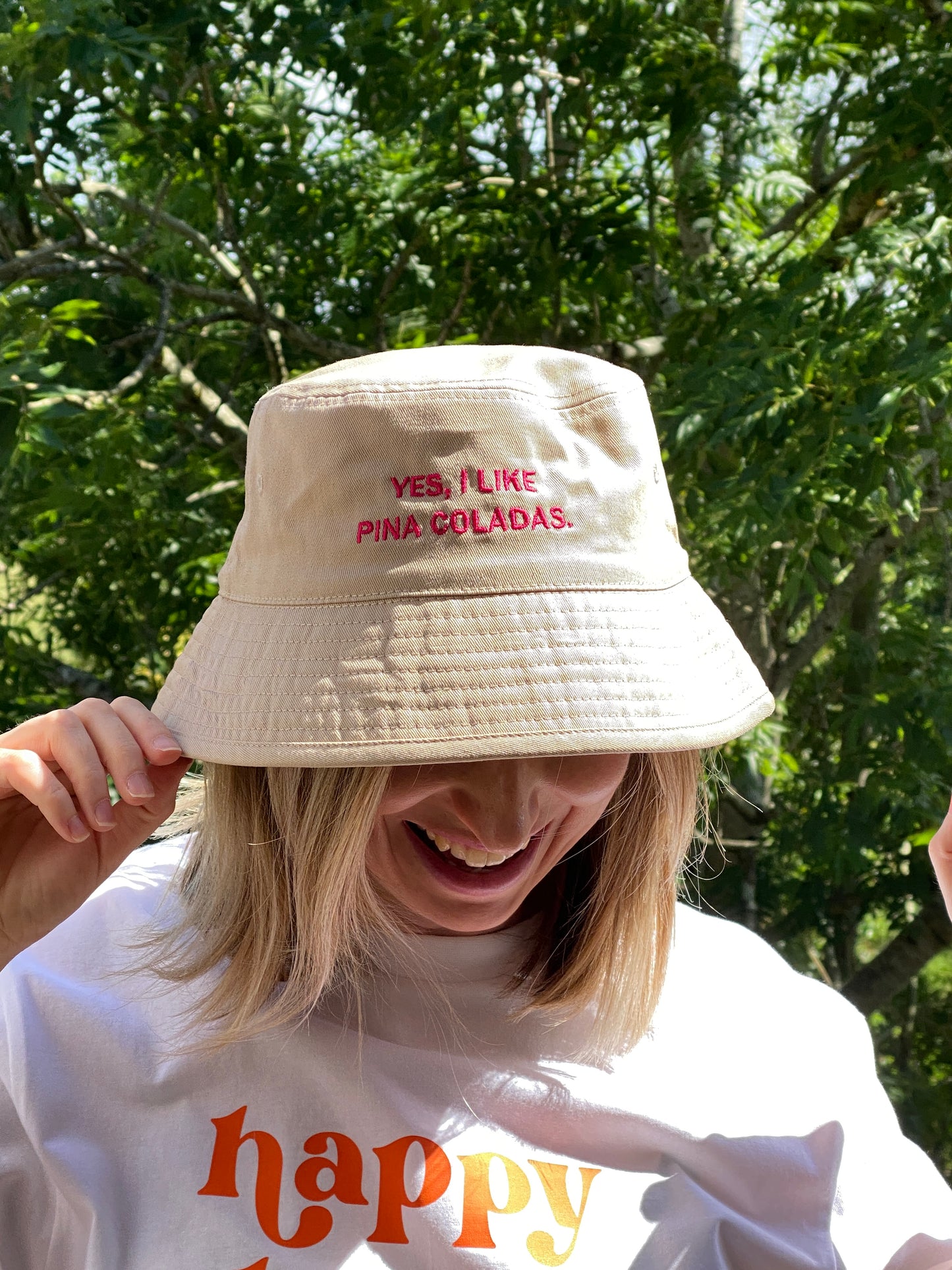 Woman wearing a cream bucket hat embroidered with the slogan 'Yes I like pina coladas' in hot pink