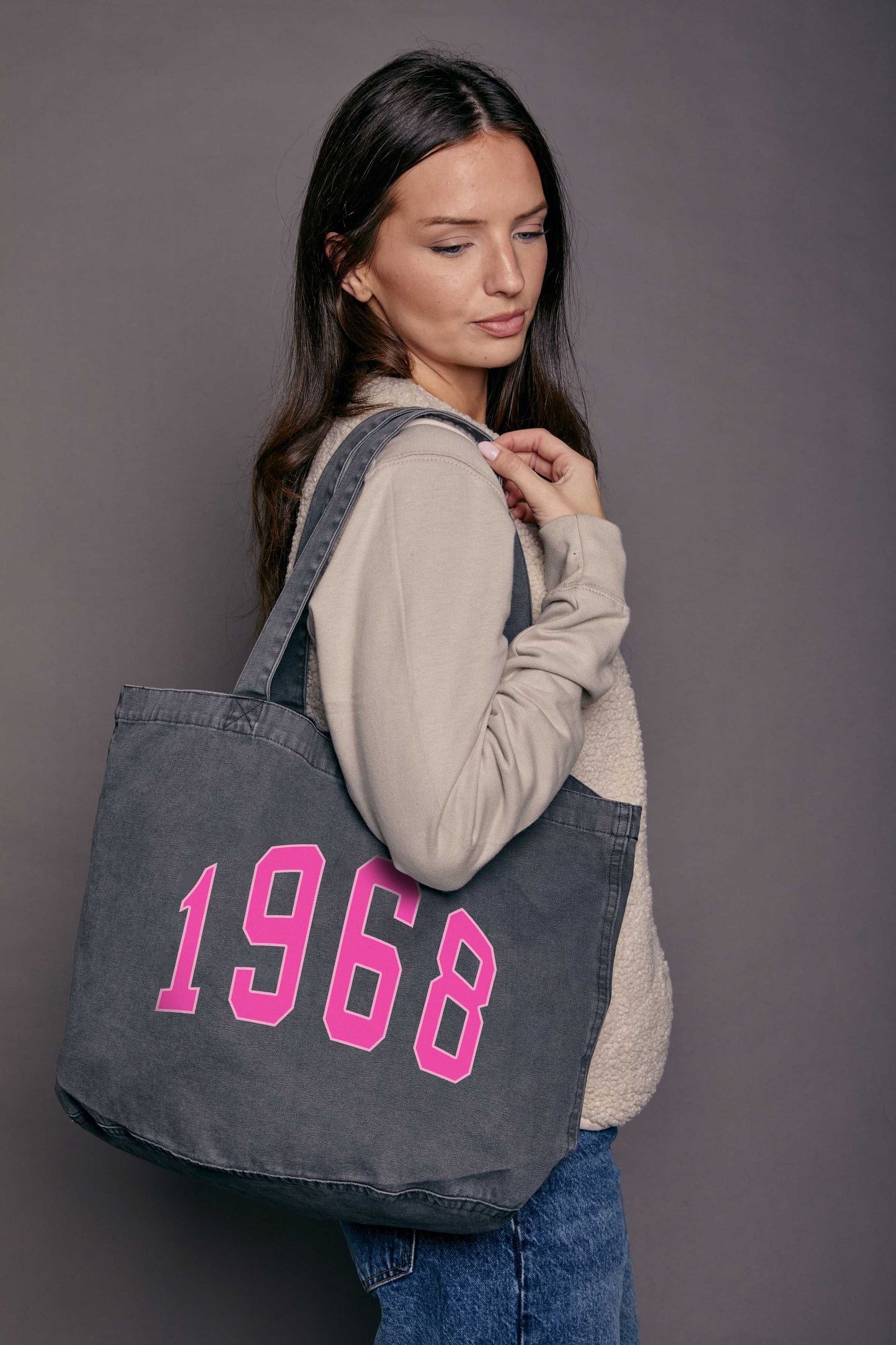 Woman holding a black denim tote bag with '1968' printed on it in pink against a grey background