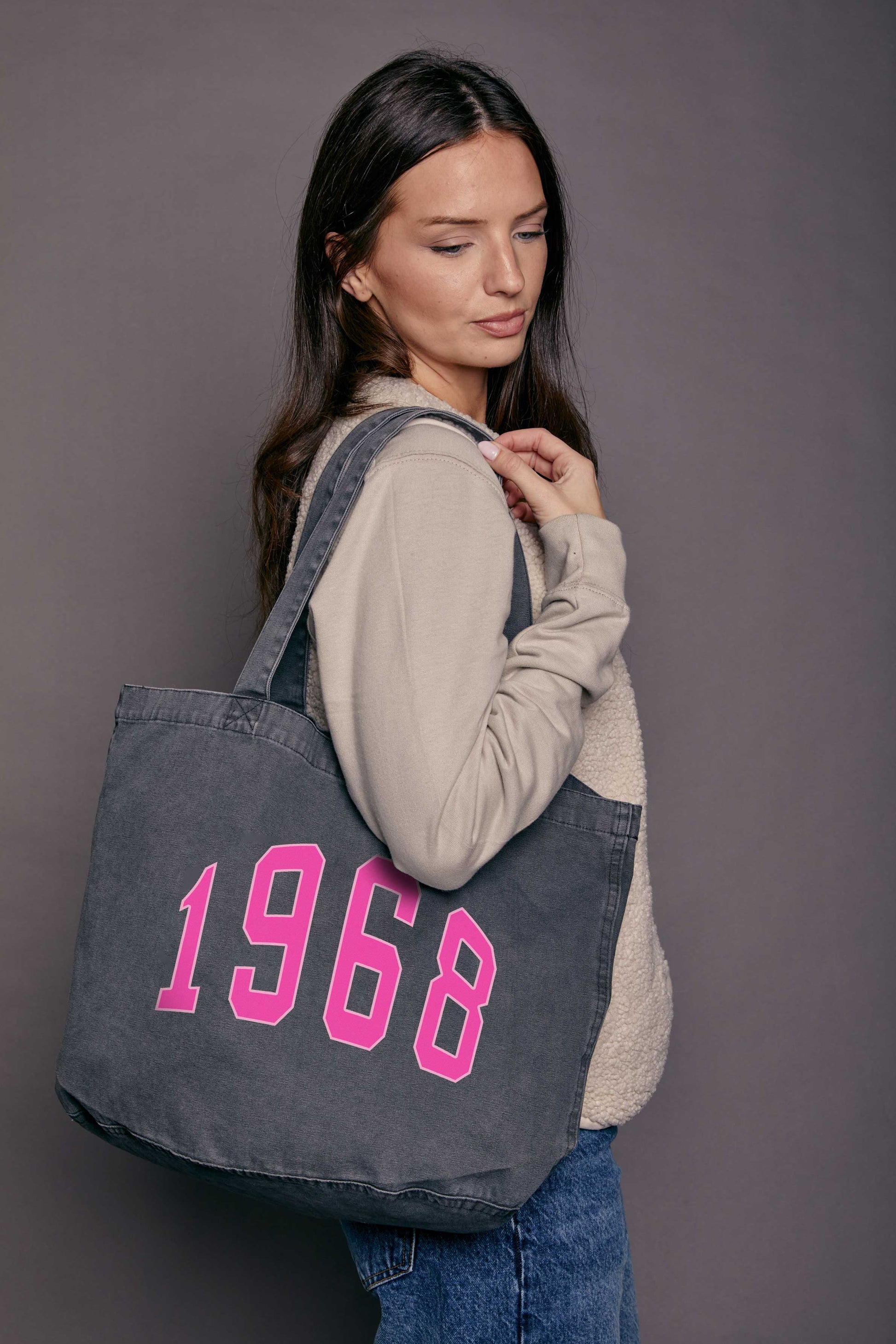 Woman holding a black denim tote bag with '1968' printed on it in pink against a grey background