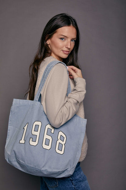 Woman carrying a blue denim tote bag with '1968' printed on it against a grey background
