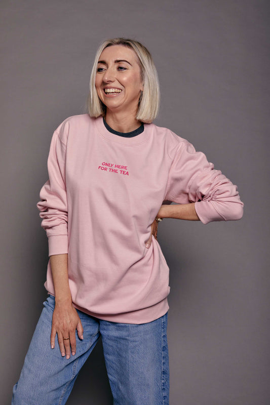 Woman wearing a pink sweatshirt with hot pink embroidered text 'Only here for the tea', standing against a grey background