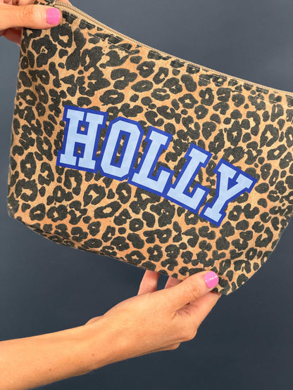 Leopard print pouch bag with 'HOLLY' printed slogan held by a hand against a dark background