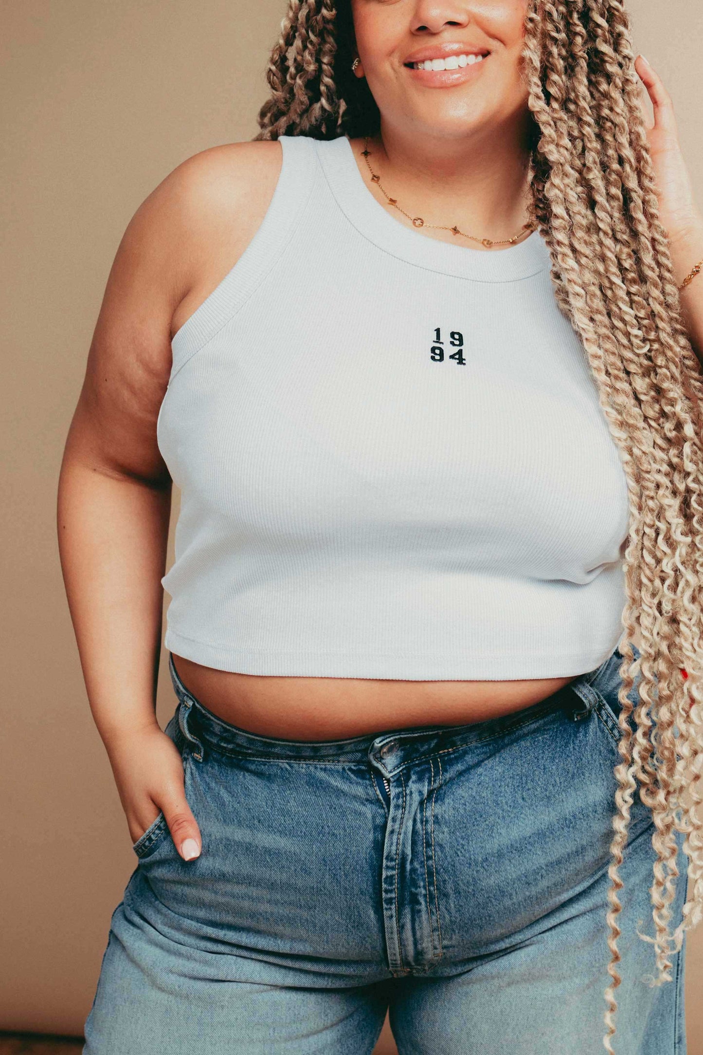 Person wearing a white  tank top with '1984' embroidery and high-waisted jeans against a beige background