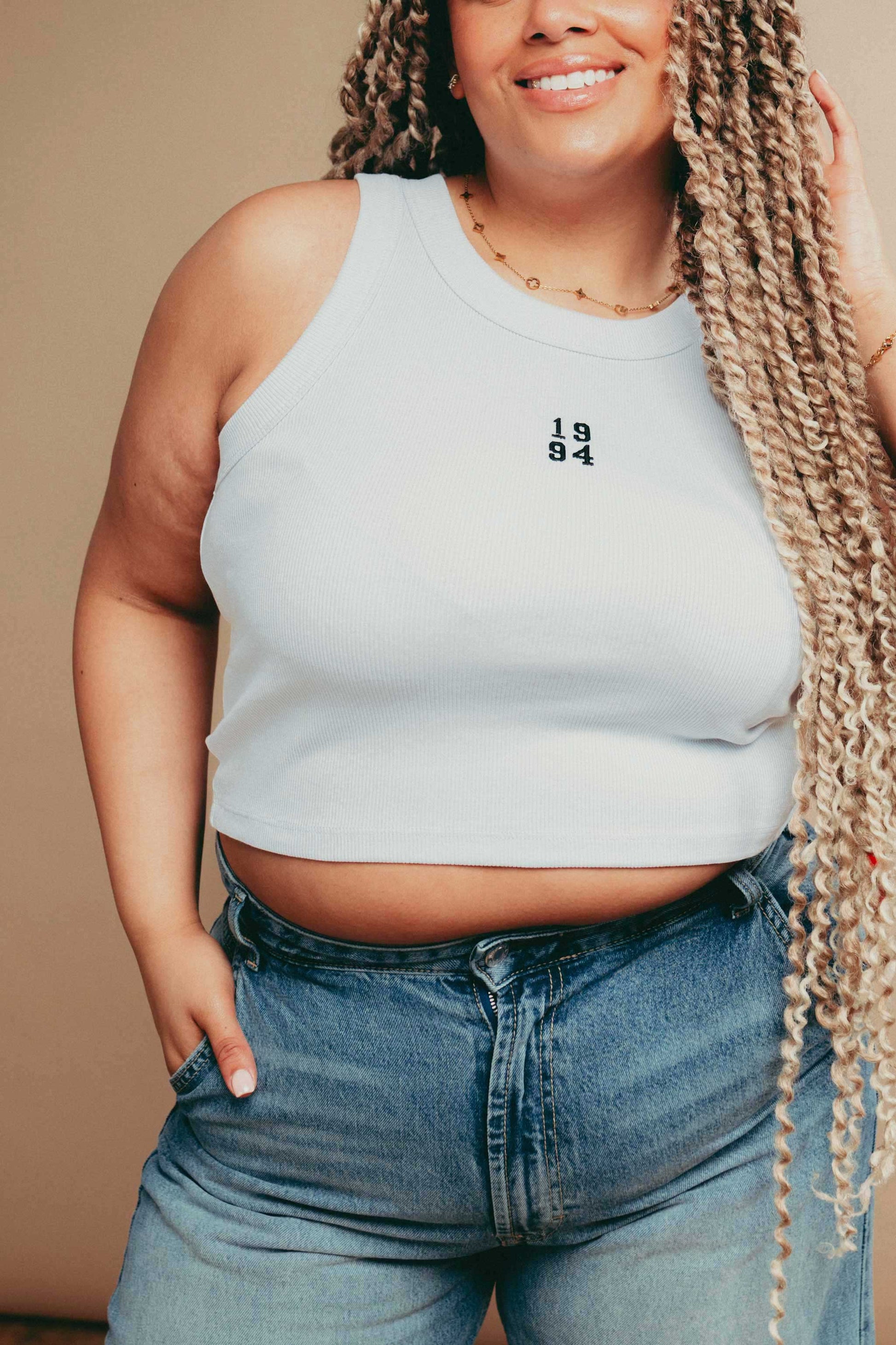 Person wearing a white  tank top with '1984' embroidery and high-waisted jeans against a beige background