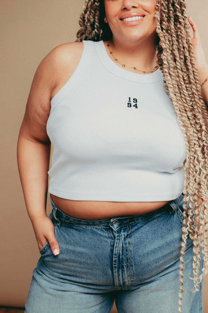 Person wearing a white  tank top with '1984' embroidery and high-waisted jeans against a beige background