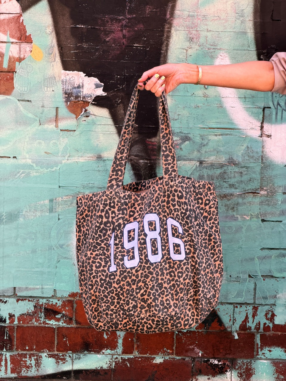 Leopard print tote bag with '1986' text held by a hand against a textured wall.