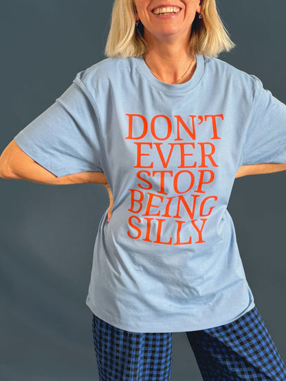 A woman wearing a light blue graphic t-shirt with the phrase 'DON'T EVER STOP BEING SILLY' printed in orange text, paired with blue and black checkered trousers, against a dark gray background.