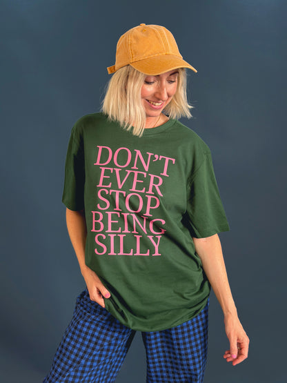 A woman wearing an oversized dark green t-shirt with the text 'DON'T EVER STOP BEING SILLY' printed in pink, paired with a mustard yellow baseball cap and blue plaid barrel leg trousers, set against a dark slate blue background.