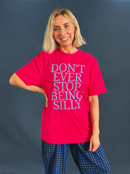 A smiling blonde woman wearing a bright pink graphic t-shirt with the text 'DON'T EVER STOP BEING SILLY' in light blue and dark pink from Rock On Ruby,  paired with blue and black checkered trousers, against a dark gray studio background.