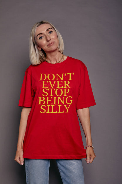 A woman with blonde hair wearing a red oversized t-shirt with yellow text that reads 'DON'T EVER STOP BEING SILLY', paired with light blue jeans, standing against a solid gray background.