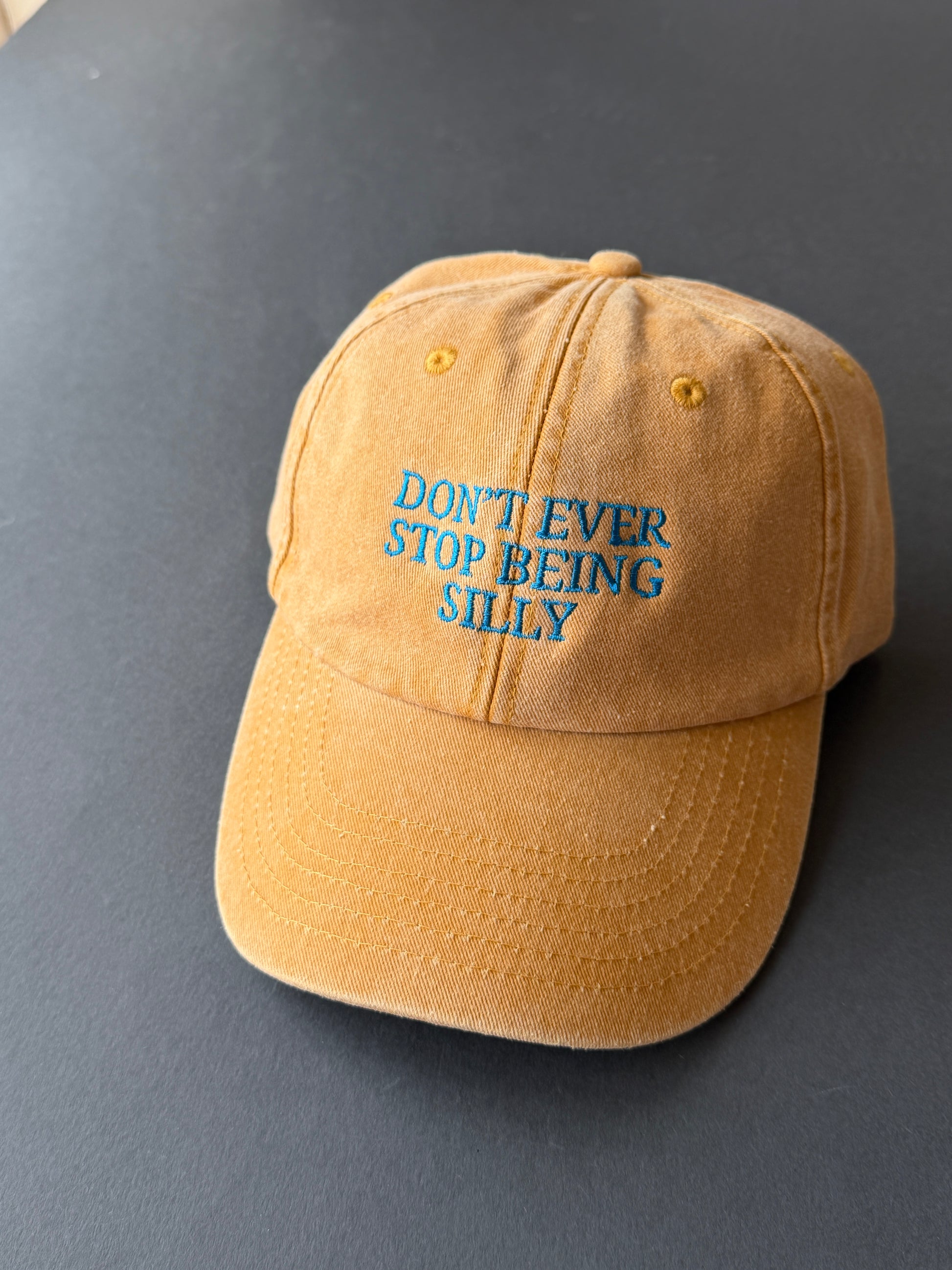 A mustard yellow baseball cap with blue embroidered text reading 'DON'T EVER STOP BEING SILLY' on a dark gray background.