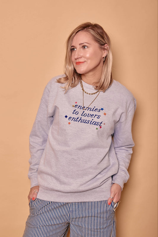 A woman wearing a light grey crewneck sweatshirt with the printed text 'enemies to lovers enthusiast' in blue, paired with blue and white striped pants, standing against a solid beige background.
