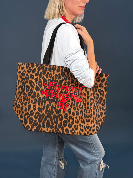 A woman wearing a white long-sleeve shirt and ripped blue jeans carries a large leopard-print tote bag with red cursive text reading 'Forever winging it'. The background is a solid dark slate blue.