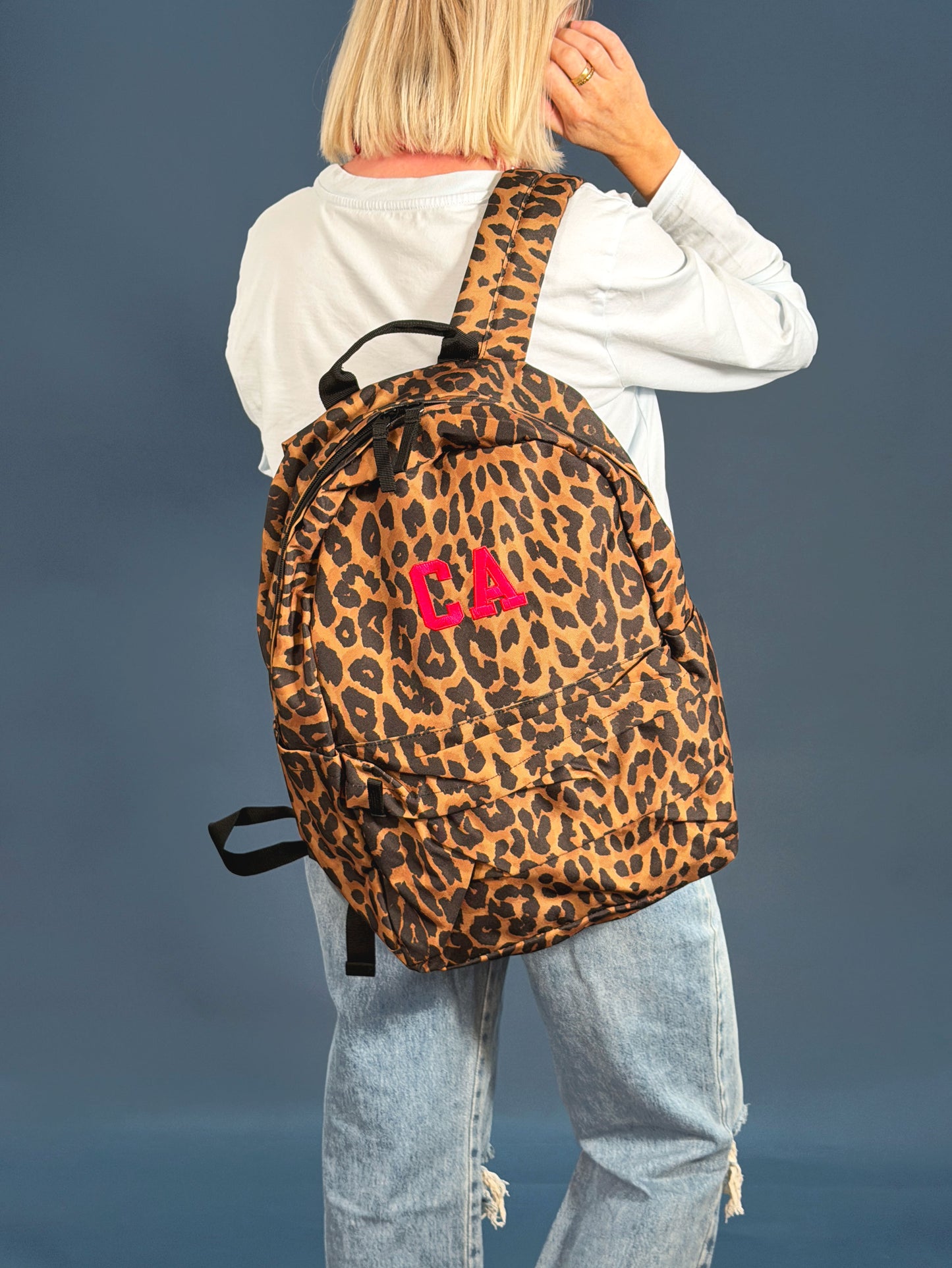 A person shown from behind wearing a leopard-print backpack with a bright pink embroidered 'CA' monogram, styled with a white long-sleeve shirt and light-wash jeans against a solid slate blue background.
