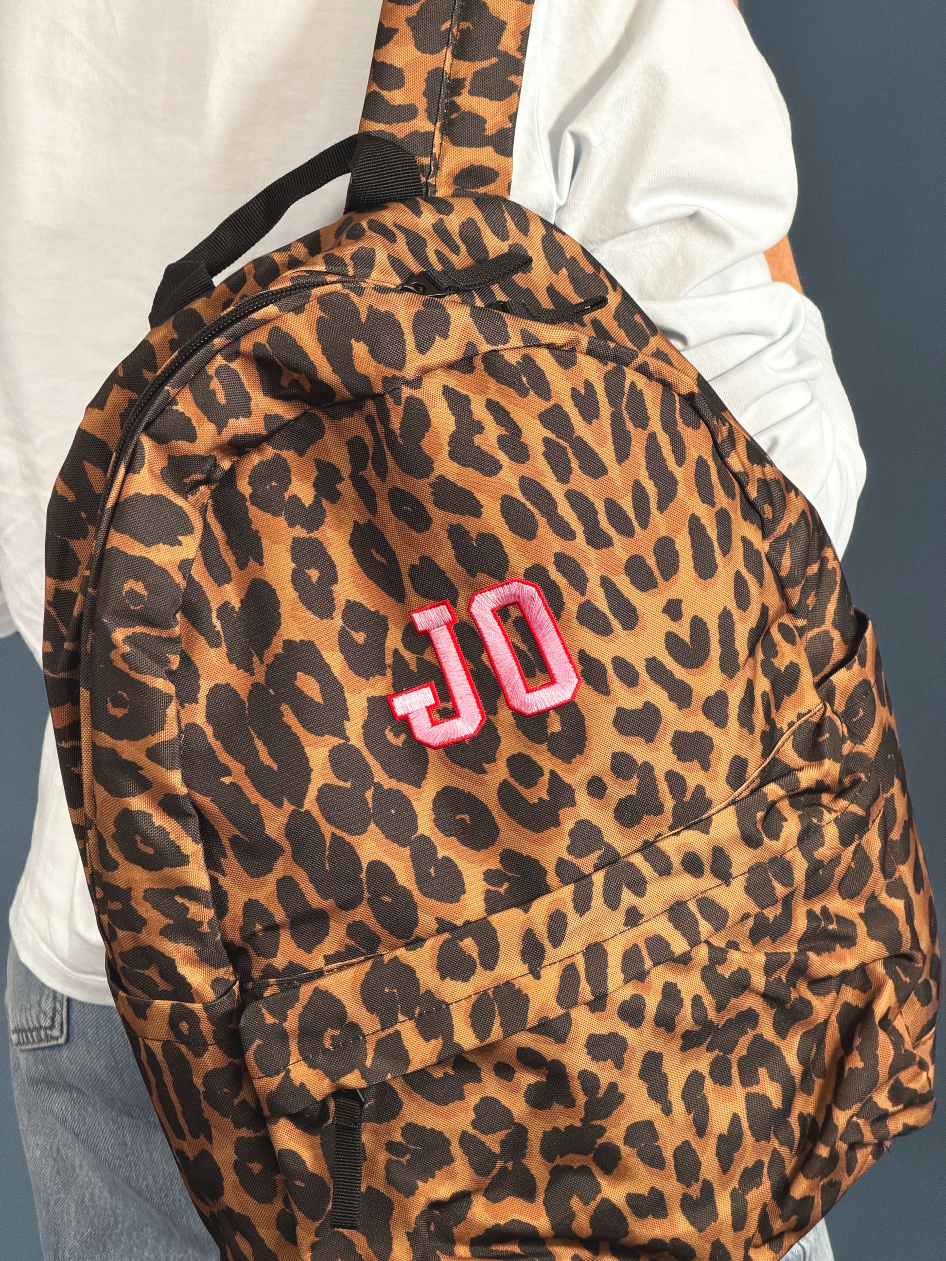 A leopard print backpack with a pink embroidered 'JO' monogram, shown being worn by a person in a white shirt and jeans.