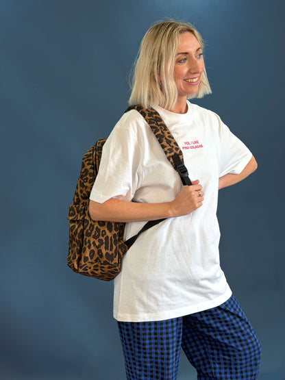 A woman with blonde hair is shown from the waist up, wearing a white t-shirt, blue and black checkered pants, and carrying a leopard print backpack against a solid dark blue background.
