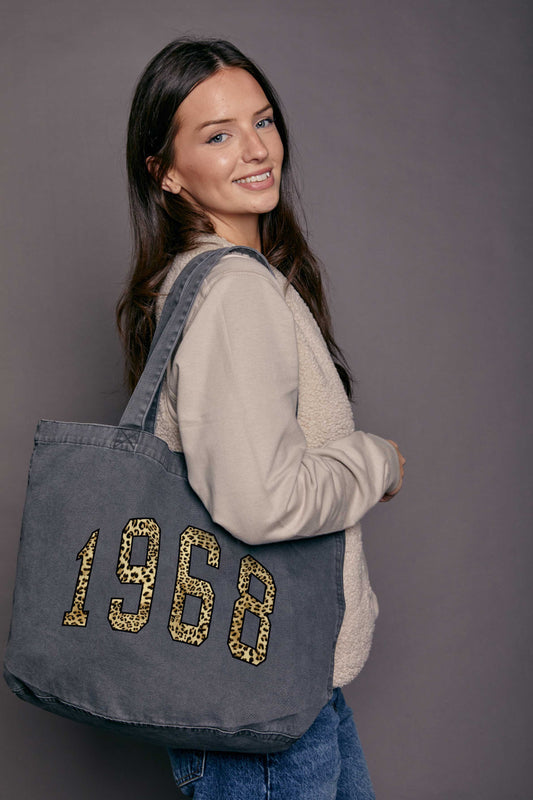 Woman carrying a faded black denim tote bag with '1968' printed on it in leopard print text