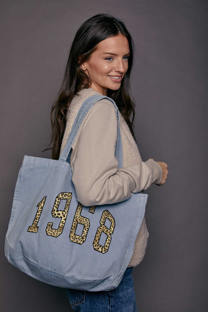 Woman carrying a blue denim tote bag with leopard print '1969' printed on it against a grey background