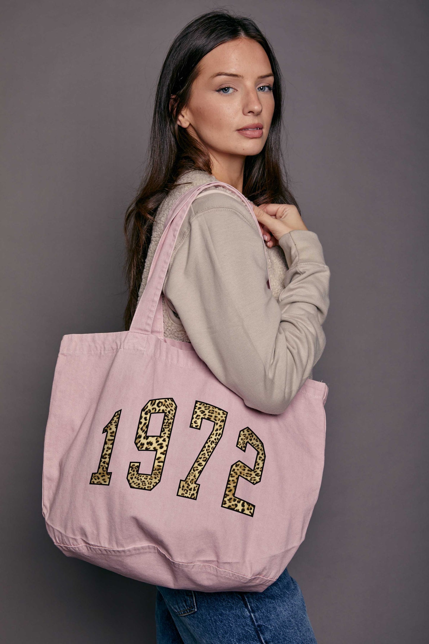 Woman holding a denim pink tote bag with '1972' printed on it against a grey background
