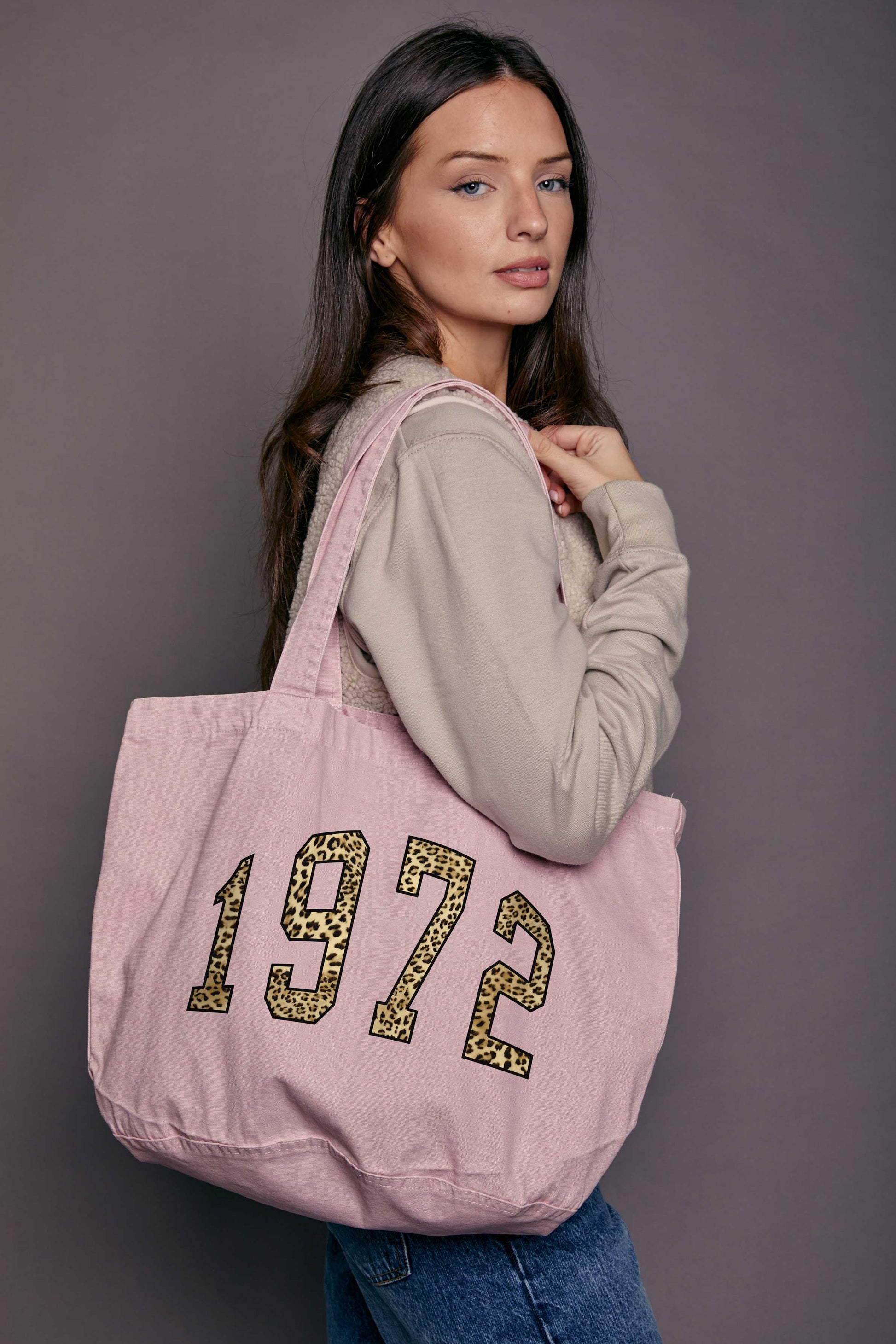 Woman holding a denim pink tote bag with '1972' printed on it against a grey background