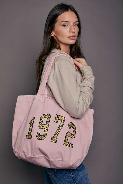 Woman holding a denim pink tote bag with '1972' printed on it against a grey background