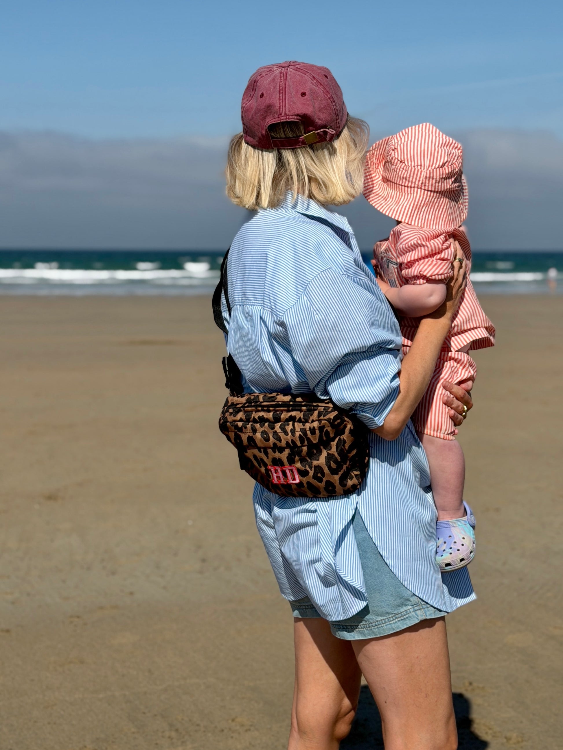 A mum and child standing on a sandy beach, viewed from behind. The woman wears a light blue striped shirt, denim shorts, a dusty rose cap, and carries a leopard-print crossbody bag with pink 'HD' lettering.